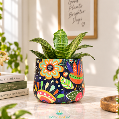 A Floral Doodle tumbler metal planter with colorful floral doodle artwork and green indoor plant styled in a modern home. Planter has a dark navy blue background and vibrant floral and leaf patterns in orange, yellow, green, and red. The pot is styled on a marble tabletop next to a stack of books, with a framed print on the wall behind it.