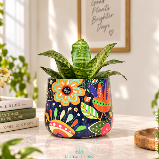 A Floral Doodle tumbler metal planter with colorful floral doodle artwork and green indoor plant styled in a modern home. Planter has a dark navy blue background and vibrant floral and leaf patterns in orange, yellow, green, and red. The pot is styled on a marble tabletop next to a stack of books, with a framed print on the wall behind it.
