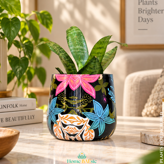 A Floral Fantasy Tumbler decorative metal planter with a black base and colorful floral illustrations, holding a snake plant on a marble table in a styled living room setting in a modern home. 