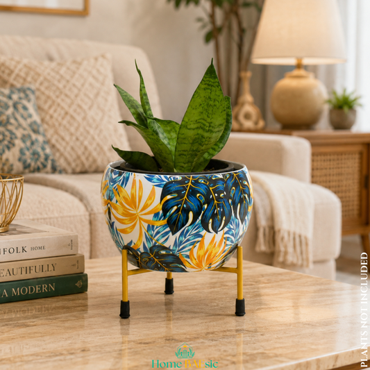 Monstera Vibes decorative metal planter with colorful blue, yellow and white tropical leaf design, displayed on a gold metal stand with a snake plant in a stylish living room.