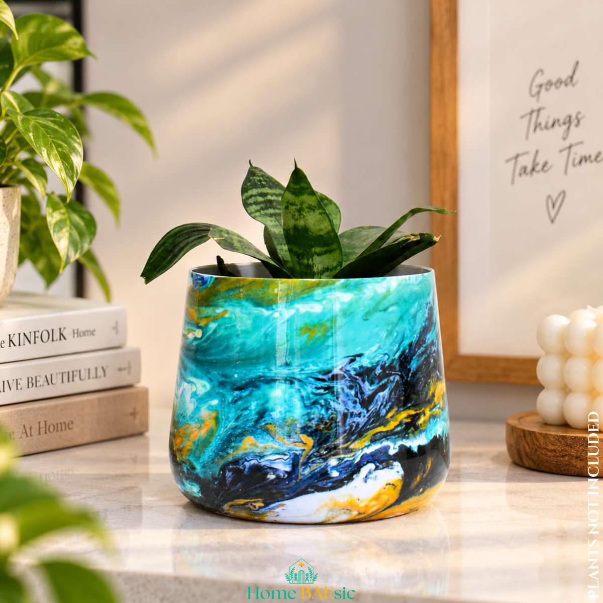 An Oceanic Turquoise tumbler planter with snake plant styled on a modern home console planter with a vibrant turquoise, blue, black, and gold marbled pattern, holding a small green snake plant, styled on a marble countertop with books and a candle in a warm, sunlit room.