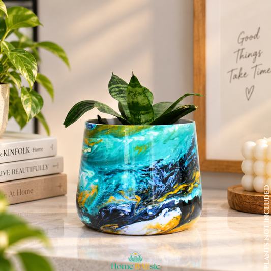 An Oceanic Turquoise tumbler planter with snake plant styled on a modern home console planter with a vibrant turquoise, blue, black, and gold marbled pattern, holding a small green snake plant, styled on a marble countertop with books and a candle in a warm, sunlit room.